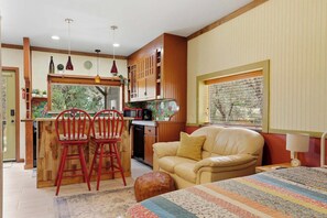 A compact kitchen with a central island, a dining area with two red chairs, a beige sofa, and a window with blinds. - Charming 1BR Studio with Full Kitchen (Prescott)