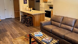 Ein Wohnzimmer mit braunem Ledersofa, einem Couchtisch aus Holz mit Zeitschriften, einem gemusterten Teppich und einer Küchenzeile mit Holzschränken und einem weißen Kühlschrank.
