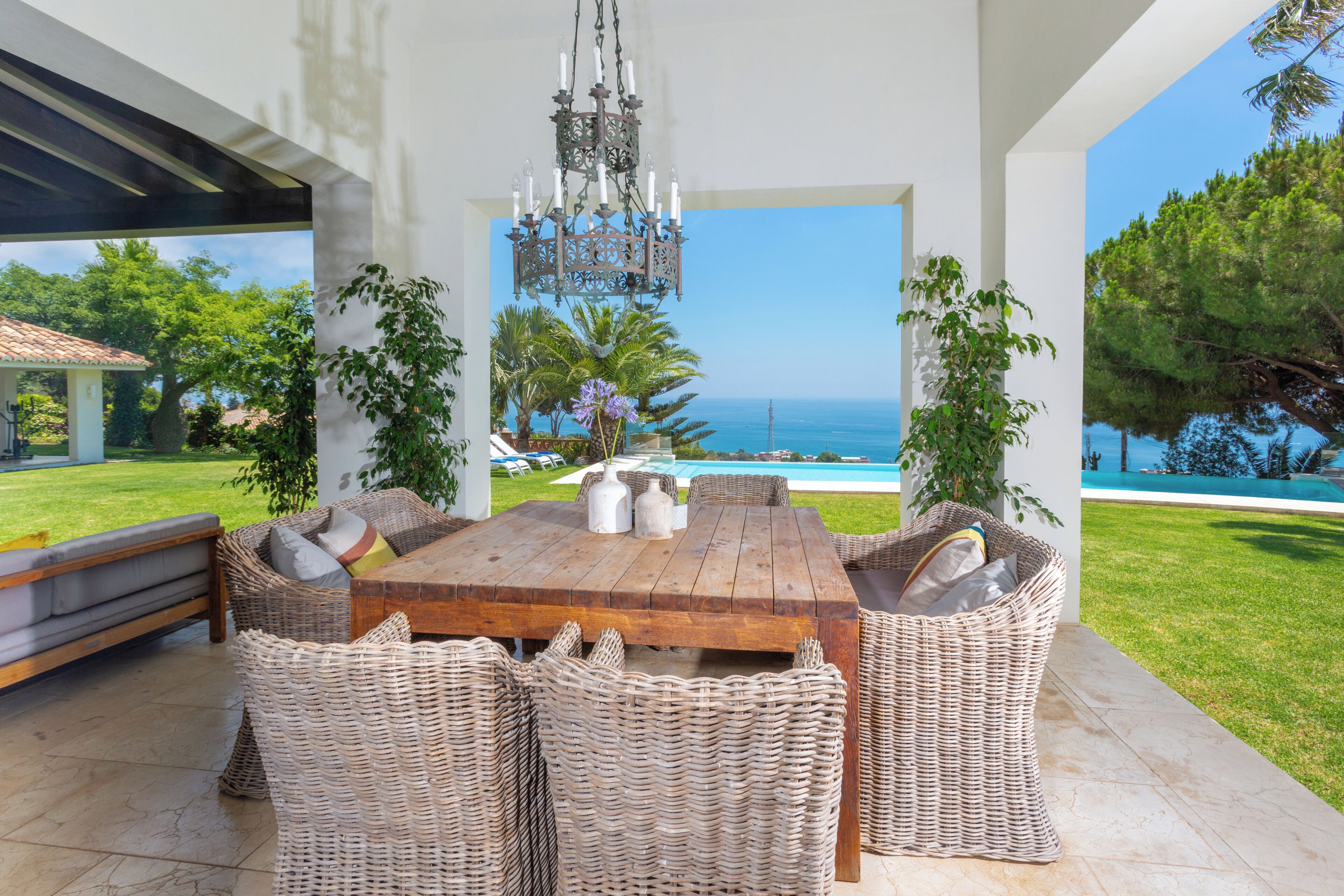 Un coin repas avec une table en bois et des chaises en osier, donnant sur une piscine et offrant une vue sur l’océan.