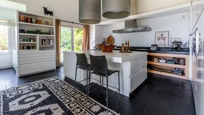 Eine moderne Küche mit einer zentralen Kochinsel, Barhocker, Pendelleuchten und Blick auf Grünflächen.