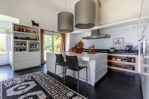 Eine moderne Küche mit einer zentralen Kochinsel, Barhocker, Pendelleuchten und Blick auf Grünflächen.
