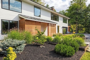 A modern two-story house with a sloped roof, large windows, and a well-maintained garden with various shrubs and a small tree. - Charming 2BR Hanover Apt | Walk to Dartmouth (Hanover)