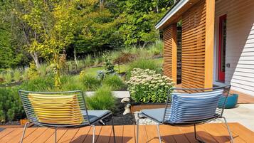 Une terrasse avec deux chaises et une petite table, entourée d’un jardin où poussent différentes plantes, et une structure en bois en arrière-plan.