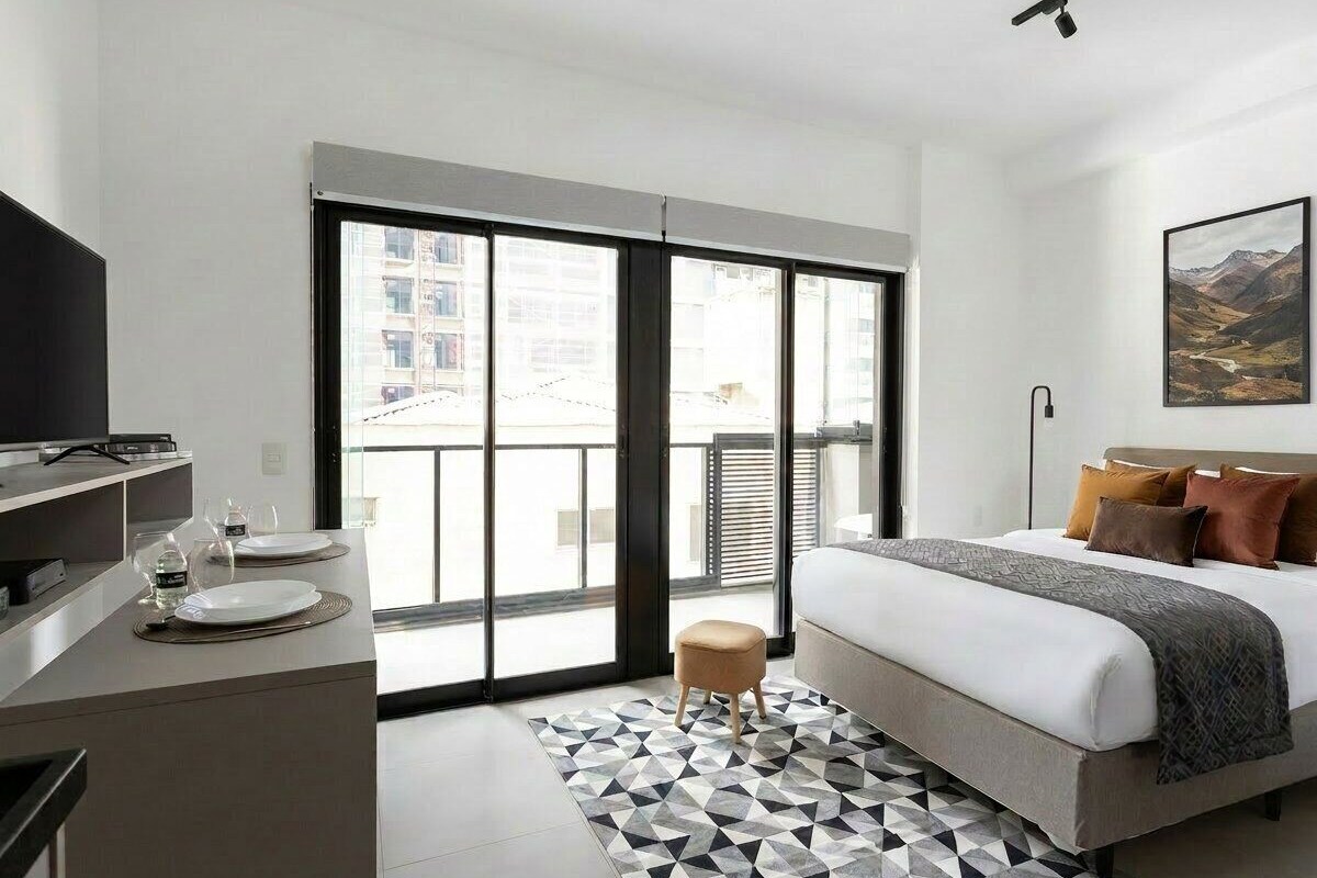A modern hotel room with a bed, a TV, a dining area with plates and glasses, and a large window with a city view.
