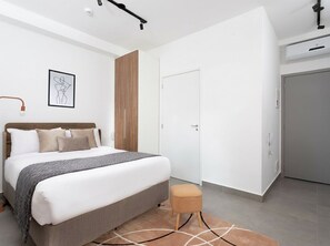 A modern bedroom with a large bed, a bedside table, a small stool, and a door. - VN Capote Valente (São Paulo)