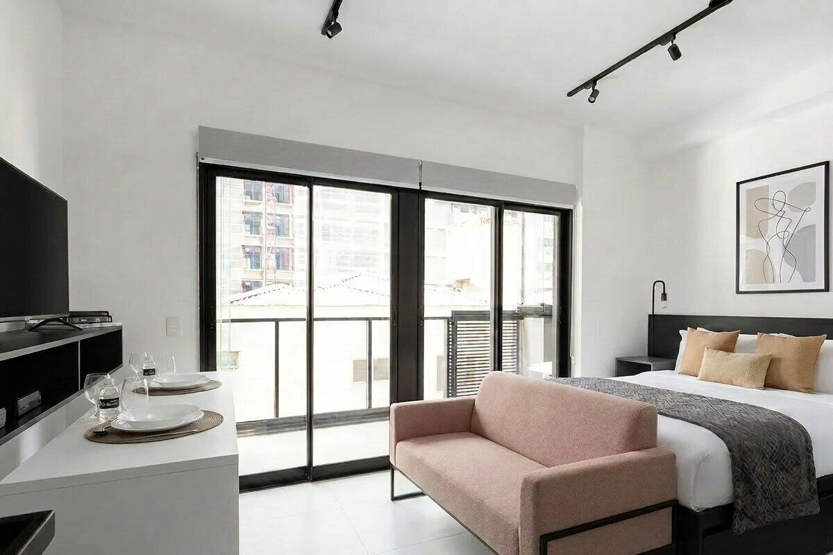 A modern hotel room with a bed, a sofa, a desk with a flat-screen TV, and a large window with a view of buildings.