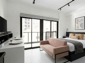Une chambre d’hôtel moderne équipée d’un lit, d’un canapé, d’un bureau avec un téléviseur à écran plat et d’une grande fenêtre donnant sur des immeubles.