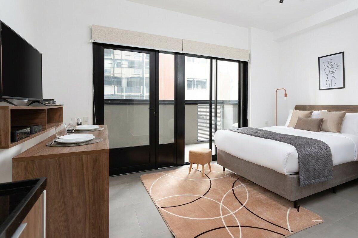 A modern hotel room with a bed, a desk with a flat-screen TV, a small stool, and a large window with a view of buildings.