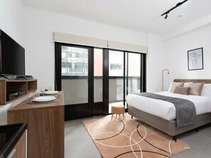 A modern hotel room with a bed, a desk with a flat-screen TV, a small stool, and a large window with a view of buildings. - VN Capote Valente (São Paulo)