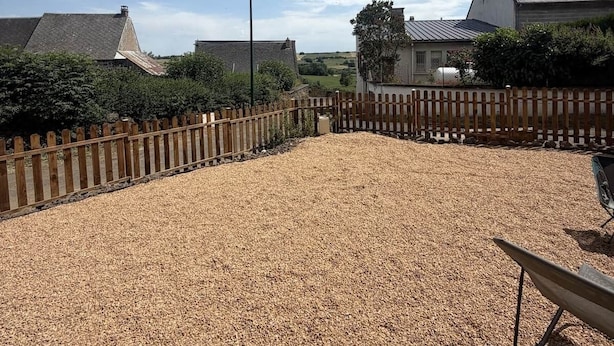 Un espacio al aire libre cubierto de grava, con una valla de madera, un banco y casas al fondo.
