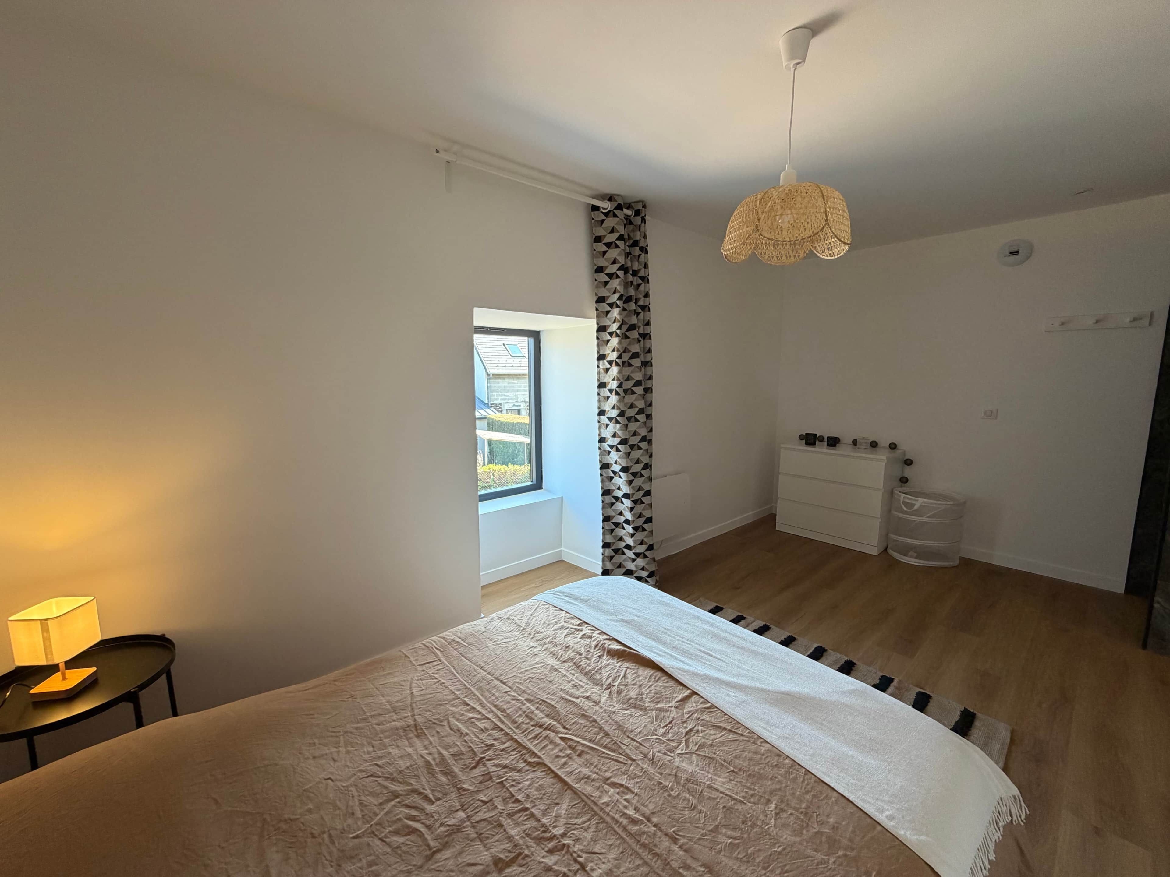 Un dormitorio con cama, mesita de noche con lámpara, armario, lámpara colgante y ventana con vistas al exterior.