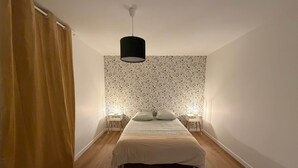 Un dormitorio con una cama, dos mesitas de noche con lámparas, una lámpara colgante y una cortina a la izquierda.