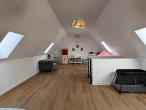 Ein geräumiges Dachzimmer mit schrägen Wänden, einem grauen Sofa, einem kleinen Tisch, einem Bücherregal und einem pinkfarbenen Bett.