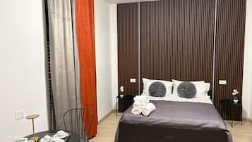 Una camera d'albergo moderna con un letto, un tavolino con una sedia, un comodino e una parete con persiane verticali.