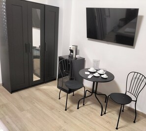 Een kamer met een garderobe, een koffieapparaat, een tafel met stoelen en een aan de muur bevestigde televisie.