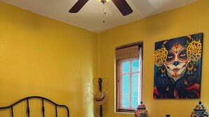 A room with a bed, a desk, a chair, a ceiling fan, a window with blinds, and a painting on the wall. - Villa Amore (Nopolo)