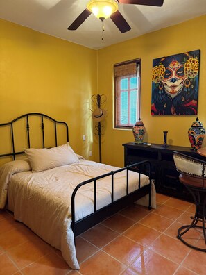 A room with a bed, a desk, a chair, a ceiling fan, a window with blinds, and a painting on the wall. - Villa Amore (Nopolo)