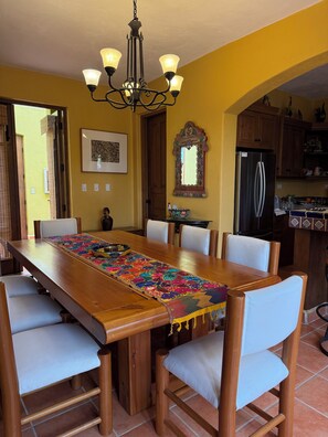 A dining area with a wooden table, white chairs, a chandelier, a mirror, and a refrigerator. - Villa Amore (Nopolo)