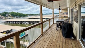 Een overdekt terras buiten met een tafel, stoelen, een plafondventilator en uitzicht op een jachthaven en strand.