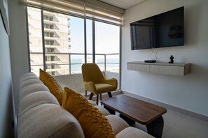 A modern living room with a sofa, armchair, and coffee table, a mounted TV, and a balcony with a view of buildings and the sea. - Golden Breeze Apartment • Beach • Seaview • Pool (Cartagena)