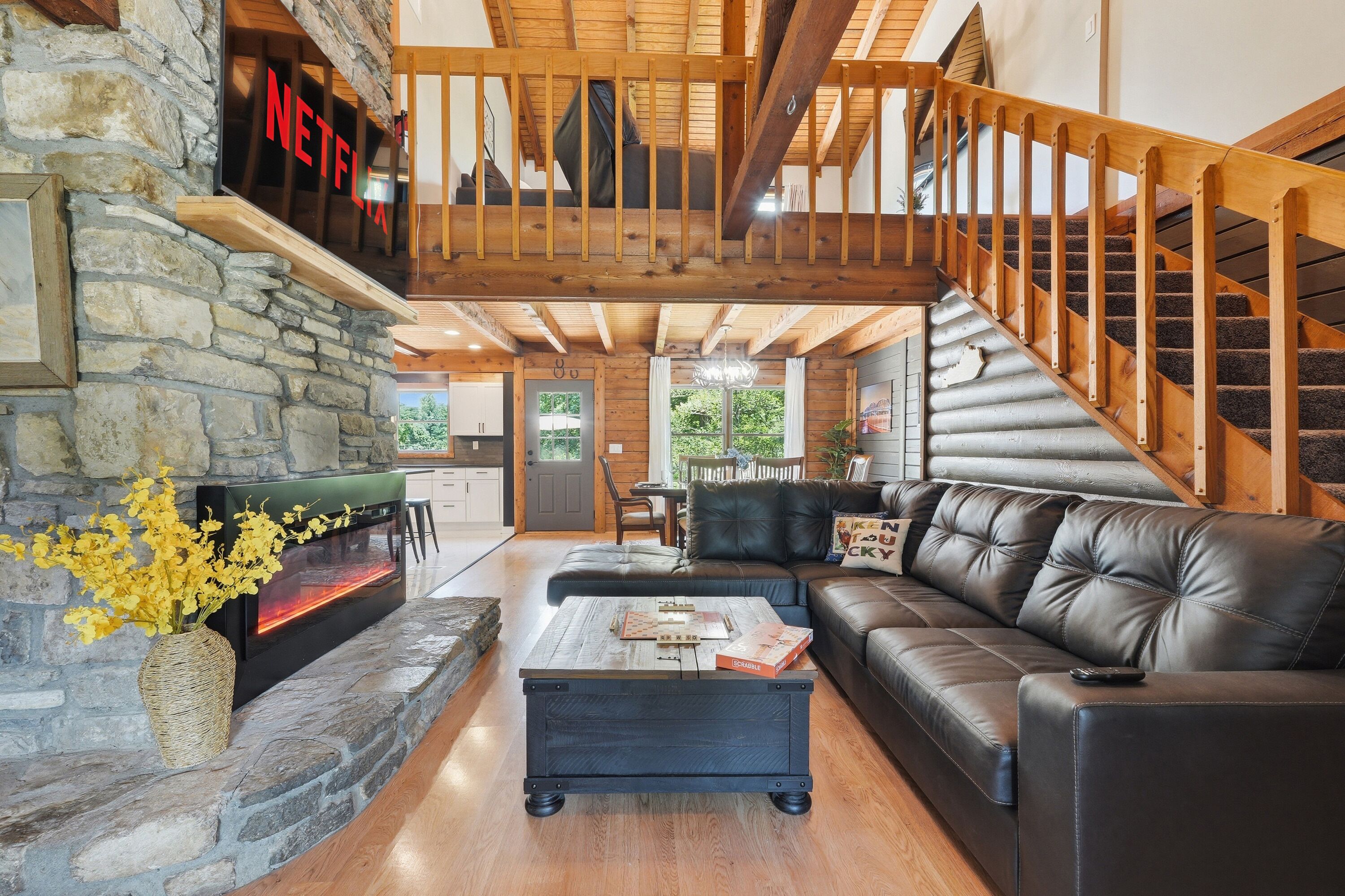 A cozy living room with a stone fireplace, a black leather sofa, and a wooden staircase.