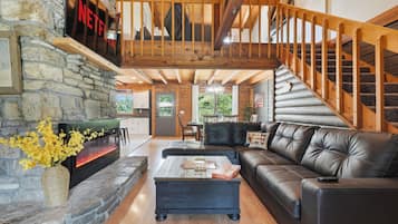 A cozy living room with a stone fireplace, a black leather sofa, and a wooden staircase.