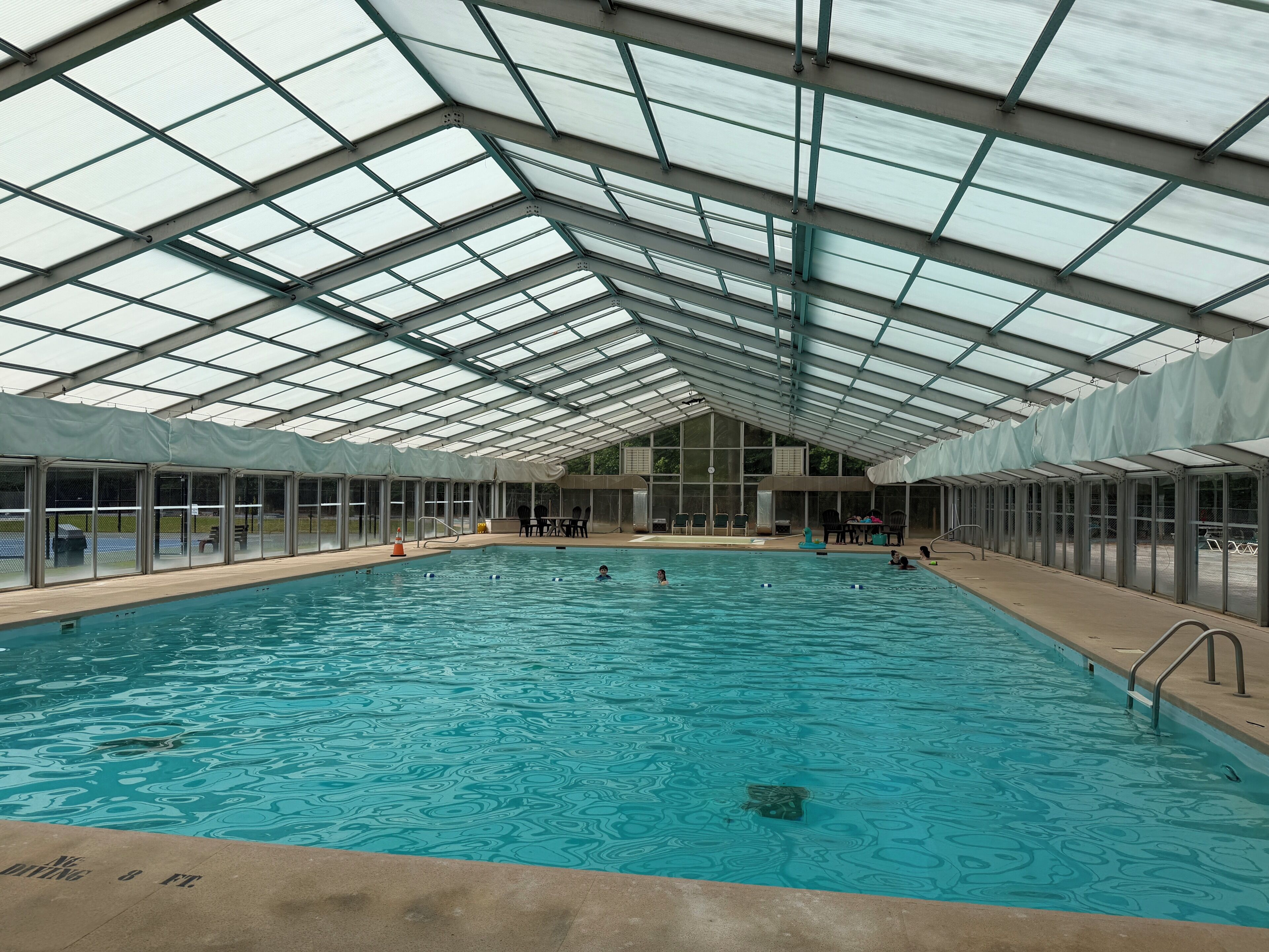 Ein Hallenbad mit hoher Decke und Glaswänden, umgeben von Stühlen und Tischen.