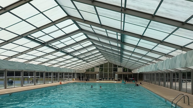 An indoor swimming pool with a high ceiling and glass panels, surrounded by chairs and tables. - Storybook Cabin-Hot Tub, Fire Pit & Tree Top Views (Ellijay)