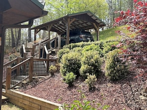 A wooden covered structure with a car parked underneath, surrounded by a landscaped garden with various shrubs and a wooden staircase leading up to it. - Storybook Cabin-Hot Tub, Fire Pit & Tree Top Views (Ellijay)