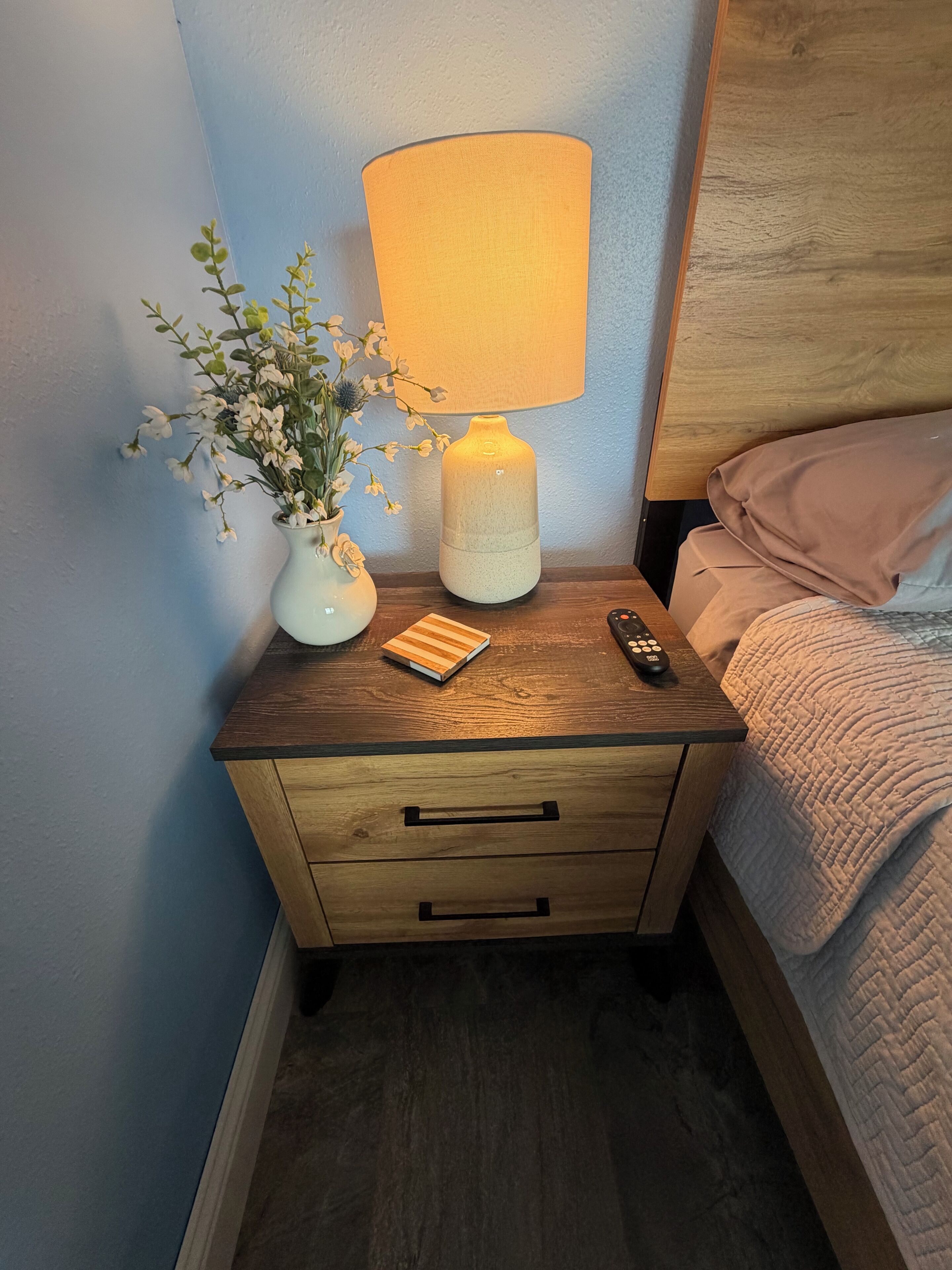 Une table de chevet en bois, avec une lampe, une t élécommande, un petit livre et un vase rempli de fleurs.