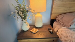 A wooden bedside table with a lamp, a remote control, a small book, and a vase with flowers. - Aeries Villas 309 A (Grafton)