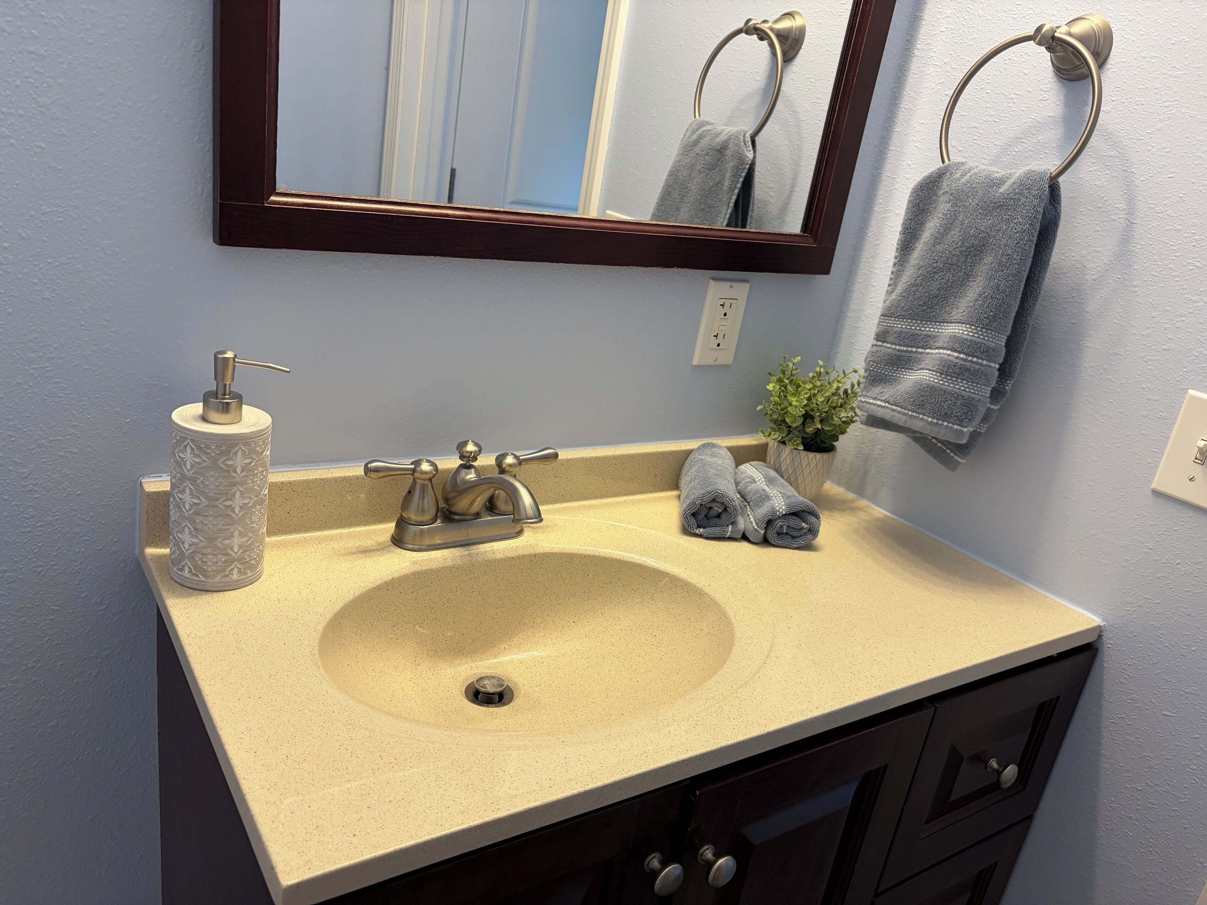 A bathroom with a beige sink, a mirror, a towel ring, a potted plant, and a towel on the counter.