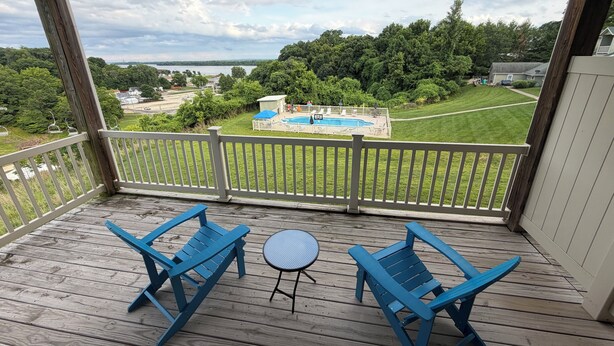 Une terrasse en bois avec deux chaises bleues et une petite table ronde, donnant sur un jardin avec une piscine et une pelouse verdoyante.