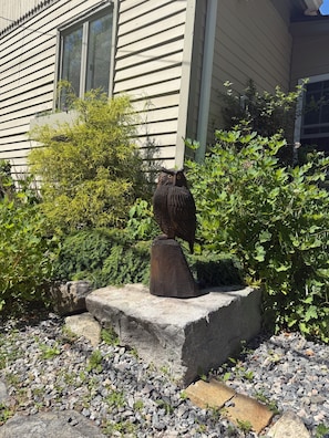 A garden with a wooden sculpture of an owl on a stone pedestal, surrounded by greenery and a house in the background.