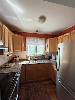 A kitchen with wooden cabinets, a granite countertop, a stainless steel refrigerator, and a window with a white valance.