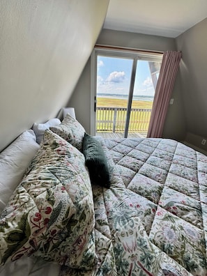 A bedroom with a large bed, floral bedding, a window with a view of a grassy area, and a balcony.