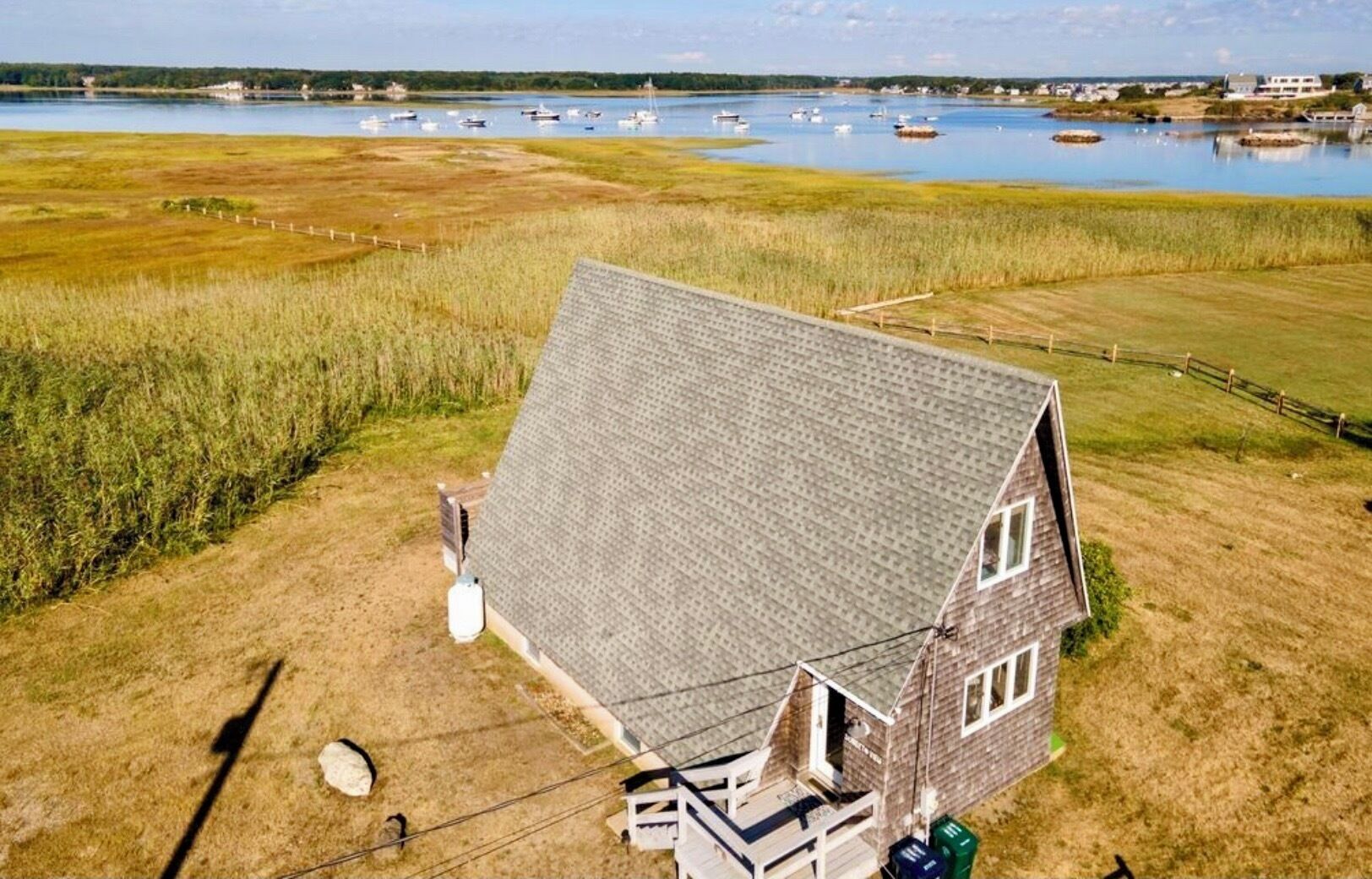 A two-story house with a sloped roof, surrounded by a grassy field and a body of water with boats.