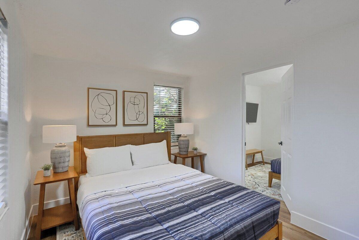 A bedroom with a bed, two bedside tables, a nightstand, a window with blinds, and a door.