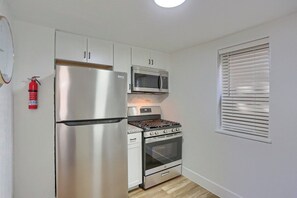 Eine kleine Küche mit Edelstahlkühlschrank, Mikrowelle und Backofen, weißen Schränken, einem Feuerlöscher und einem Fenster mit Jalousien.
