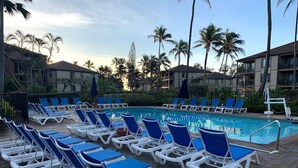 A poolside area with sun loungers and a swimming pool, surrounded by palm trees and hotel buildings.
