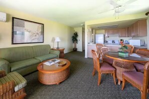 Una sala de estar con sofá, una mesita redonda, sillas de mimbre, una mesa de comedor y una cocina al fondo.