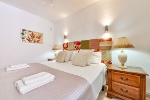 Ein ordentlich bezogenes Bett mit Kissen, ein Nachttisch mit Lampe und ein an der Wand hängendes Kunstwerk.