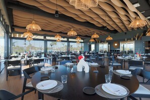 A spacious restaurant with multiple tables, hanging wicker lamps, and large windows offering a view of the city. - Brand new 3 bedroom appartement at Santa Rosalia Lake & Life Resort (MC)
