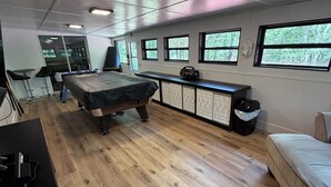 Um quarto amplo com mesa de bilhar, sofá e escrivaninha com computador. Janelas oferecem vista para as árvores.