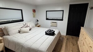 Um quarto com cama, mesas de cabeceira, cômoda, duas janelas e ar condicionado.