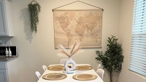 Un comedor con una mesa blanca, cuatro sillas, un mapa del mundo en la pared, una planta en maceta y una ventana con persianas.