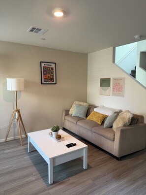 A modern living room with a white coffee table, a beige sofa with decorative pillows, a framed artwork, and a standing lamp. - Cozy Townhome, 17 min to downtown & Nellis AFB (North Las Vegas)