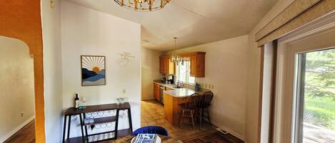 A compact kitchen with wooden cabinets, a dining area with blue chairs, and a large window.