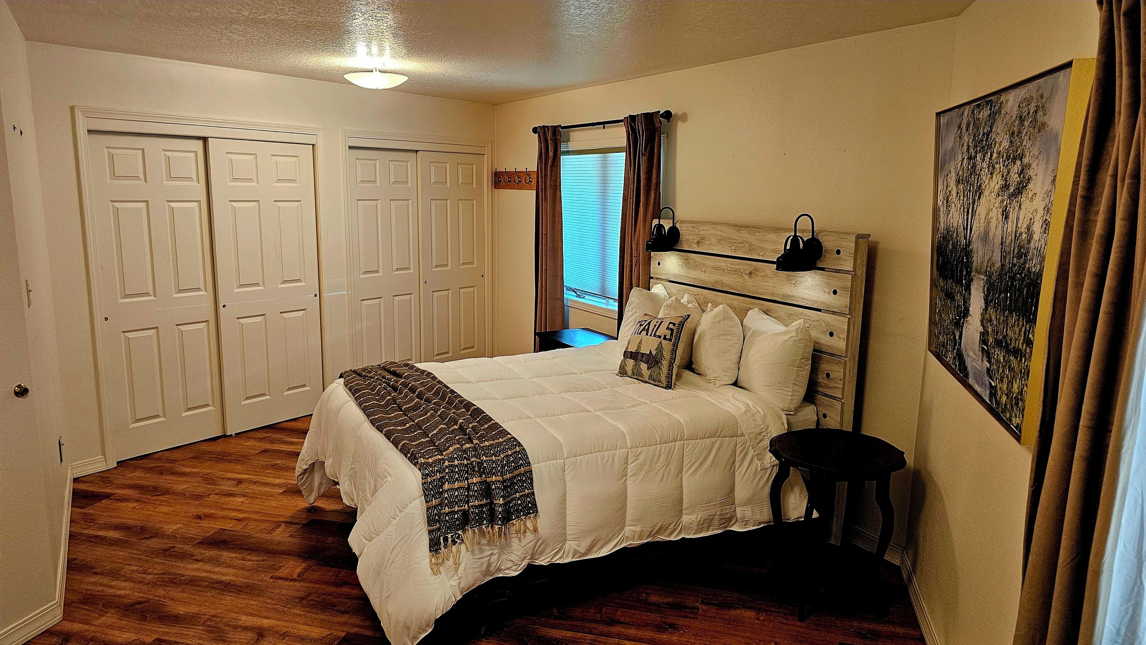 Une chambre à coucher comprenant un lit, une tête de lit, une table de chevet, une fenêtre avec des rideaux, une porte et un tableau accroché au mur.