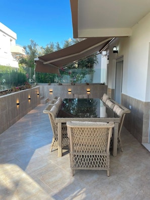Eine Terrasse im Freien mit einem Tisch aus Glas und Korbstühlen, umgeben von einer niedrigen Mauer und einem großen Sonnensegel.